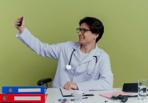 Smiling young male doctor wearing medical robe and stethoscope with glasses sitting at desk