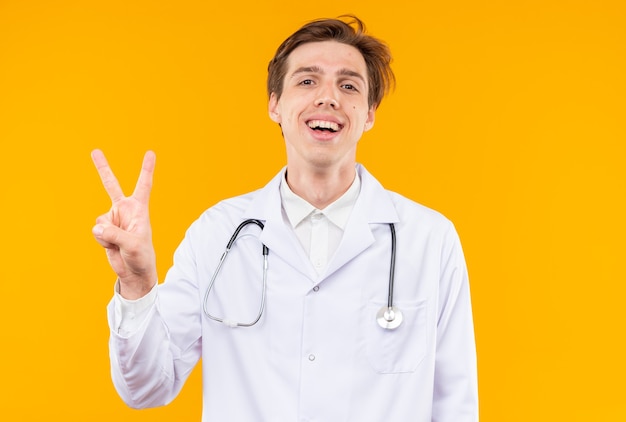 オレンジ色の壁に分離された平和のジェスチャーを示す聴診器で医療ローブを着て笑顔の若い男性医師
