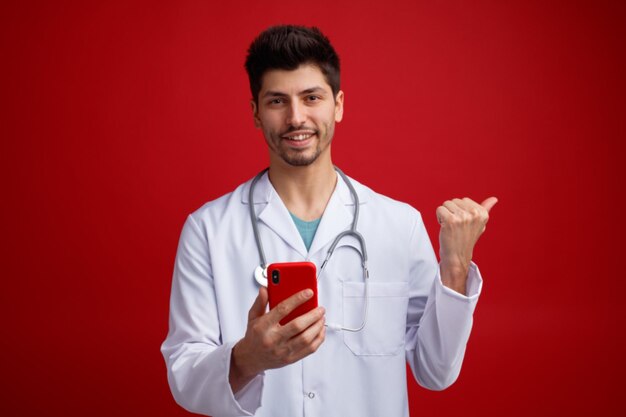 赤い背景で隔離の側を指しているカメラを見て携帯電話を保持している彼の首の周りに医療ユニフォームと聴診器を身に着けている若い男性医師の笑顔