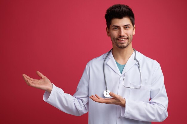 Smiling young male doctor wearing medical uniform and stethoscope around his neck looking at camera pointing to side with hands isolated on red background