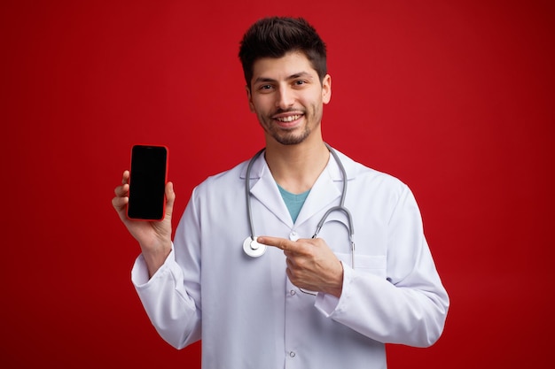 Smiling young male doctor wearing medical uniform and stethoscope around his neck looking at camera showing mobile phone to camera pointing at it isolated on red background