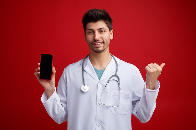 Smiling young male doctor wearing medical uniform and stethoscope around his neck looking at camera showing mobile phone pointing to side isolated on red background