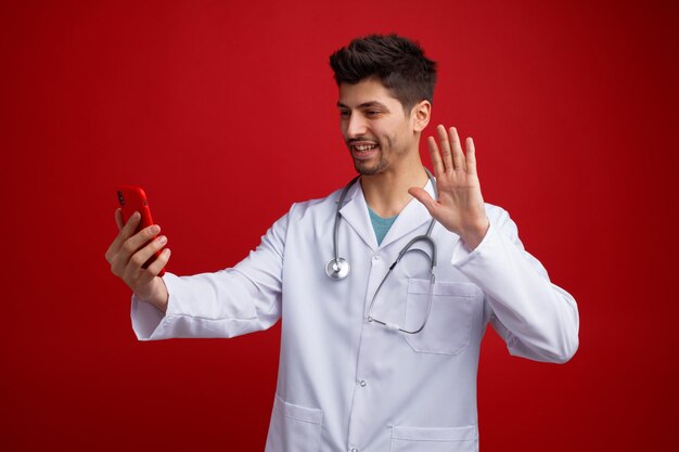 Улыбающийся молодой врач-мужчина в медицинской форме и со стетоскопом на шее растягивает мобильный телефон, разговаривая по видеосвязи, машет рукой по телефону, изолированному на красном фоне