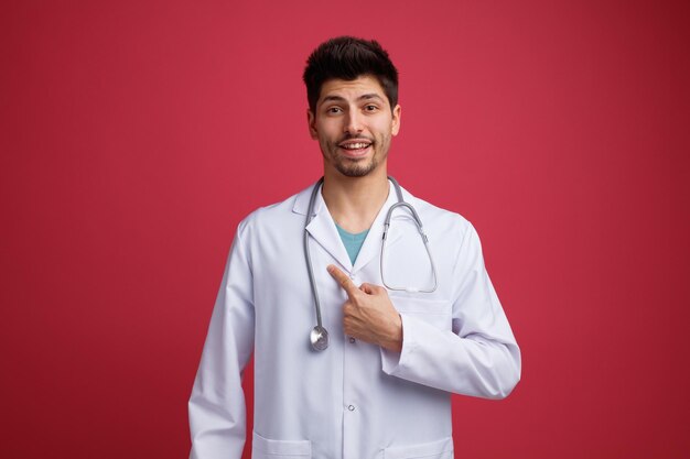 의료복을 입고 청진기를 입고 빨간 배경에 고립된 자신을 가리키는 카메라를 바라보고 있는 웃고 있는 젊은 남성 의사