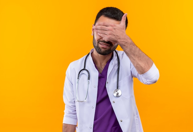 Smiling young male doctor wearing stethoscope medical gown covered eyes with hand on isolated yellow background