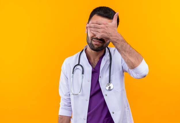 Улыбающийся молодой мужчина-врач в медицинском халате со стетоскопом закрыл глаза рукой на изолированном желтом фоне