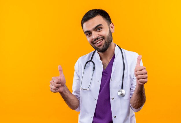 Smiling young male doctor wearing stethoscope medical gown his thumbs up on isolated yellow background