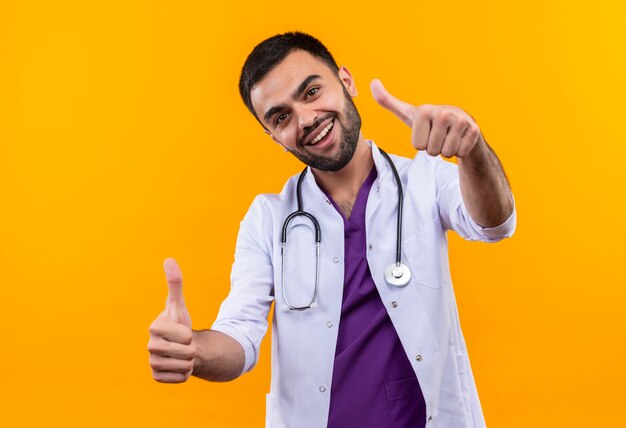 청진 기 의료 가운을 입고 웃는 젊은 남성 의사 격리 된 노란색 배경에 그의 엄지 손가락