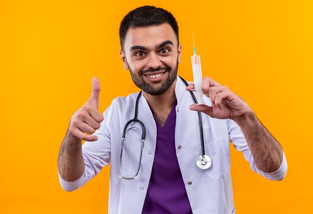 Smiling young male doctor wearing stethoscope medical gown holding syringe his thumb up on isolated yellow background