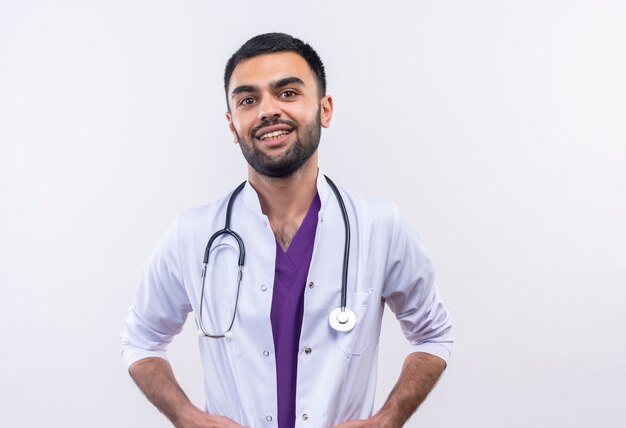 smiling young male doctor wearing stethoscope medical gown on isolated white wall