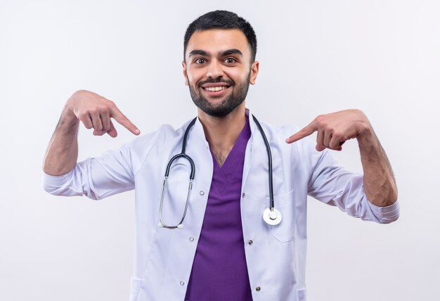 Улыбающийся молодой мужчина-врач в медицинском халате со стетоскопом указывает на себя на изолированном белом фоне