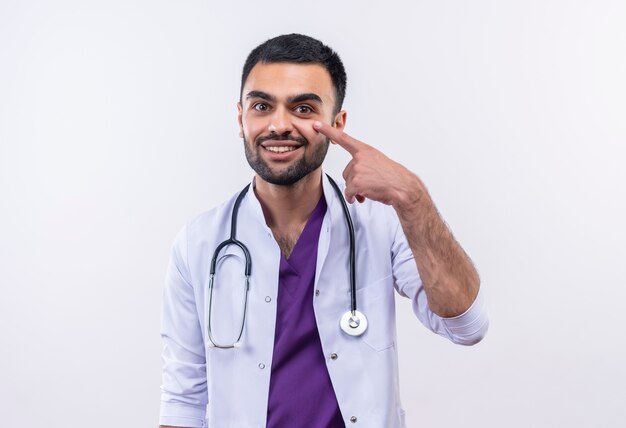 Smiling young male doctor wearing stethoscope medical gown points to his eye on isolated white background