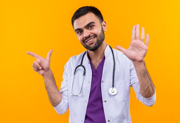 Smiling young male doctor wearing stethoscope medical gown points to side and showing stop gesture on isolated yellow background