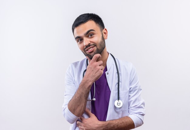 Smiling young male doctor wearing stethoscope medical gown put his hand on chin on isolated white background