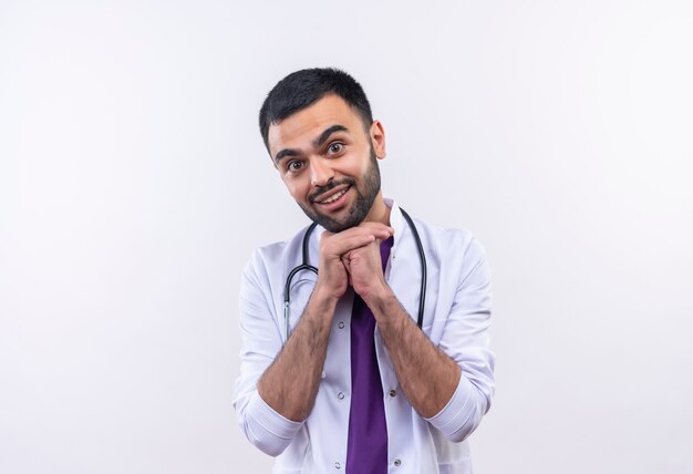 Улыбающийся молодой мужчина-врач в медицинском халате со стетоскопом положил руки на подбородок на изолированной белой стене