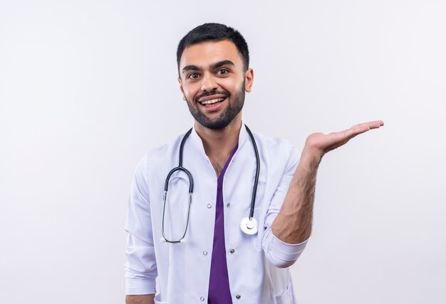 Улыбающийся молодой мужчина-врач в медицинском халате со стетоскопом, поднимающий руку на изолированном белом фоне