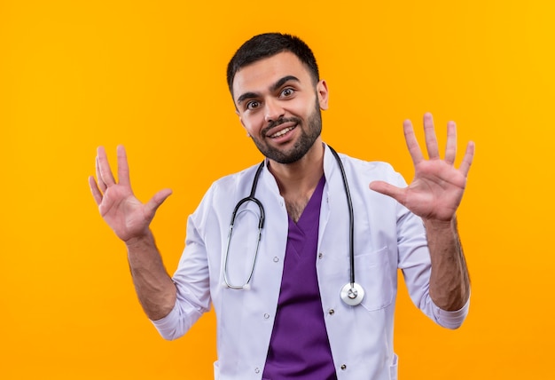 Smiling young male doctor wearing stethoscope medical gown raising hands on isolated yellow background