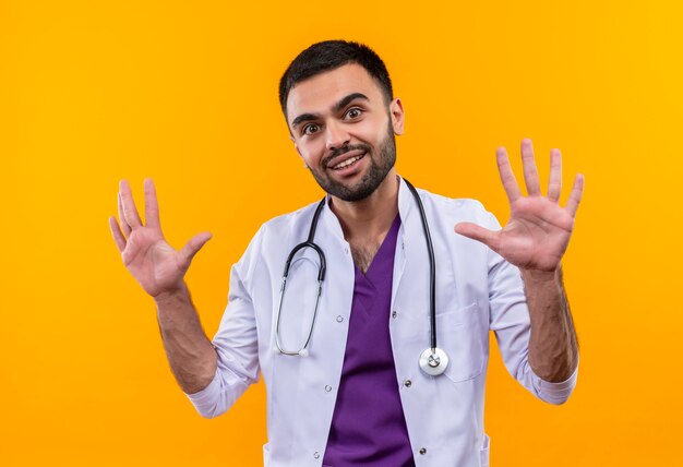 격리 된 노란색 배경에 손을 올리는 청진 기 의료 가운을 입고 웃는 젊은 남성 의사