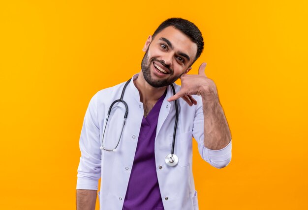 Улыбающийся молодой мужчина-врач в медицинском халате со стетоскопом показывает жест вызова на изолированном желтом фоне
