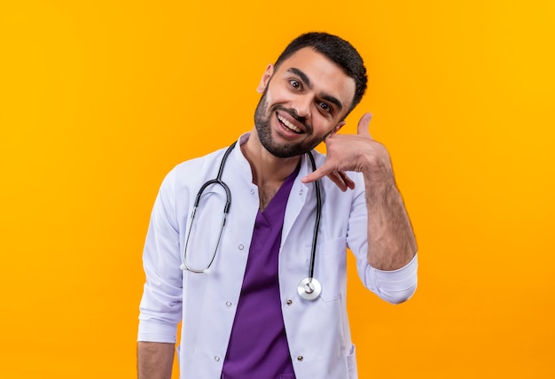 Улыбающийся молодой мужчина-врач в медицинском халате со стетоскопом показывает жест вызова на изолированном желтом фоне