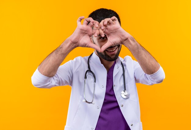 Smiling young male doctor wearing stethoscope medical gown showing heart gesture on isolated yellow background