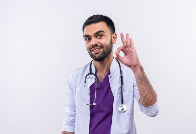 Smiling young male doctor wearing stethoscope medical gown showing okay gesture on isolated white background