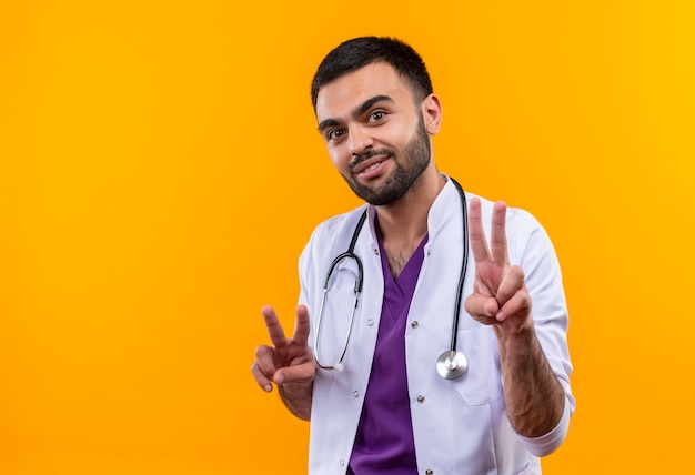 孤立した黄色の背景に平和のジェスチャーを示す聴診器の医療用ガウンを身に着けている若い男性医師の笑顔