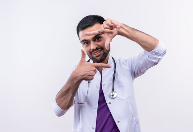격리 된 흰색 배경에 사진 제스처를 보여주는 청진 의료 가운을 입고 웃는 젊은 남성 의사