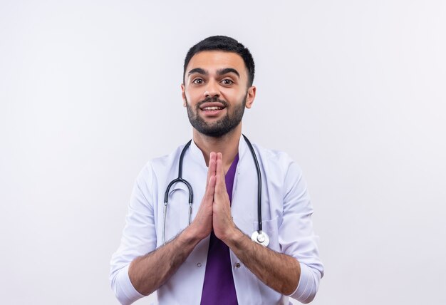 Smiling young male doctor wearing stethoscope medical gown showing pray gesture on isolated white background