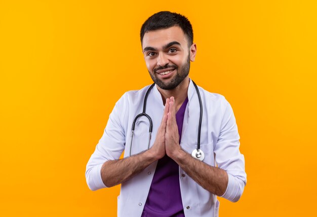 Smiling young male doctor wearing stethoscope medical gown showing pray gesture on isolated yellow background