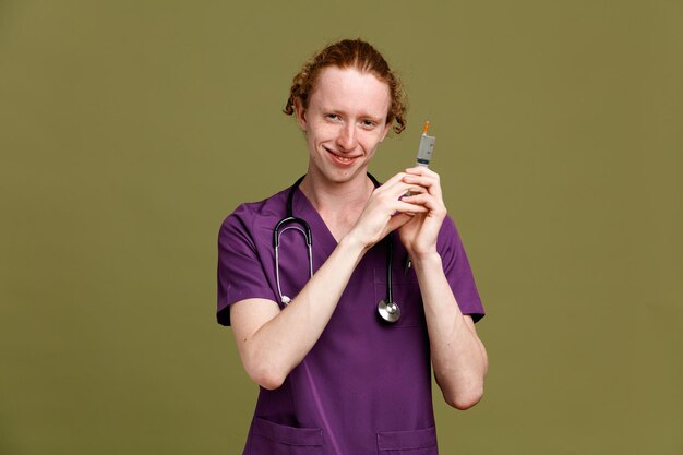 緑の背景に分離された注射器を保持している聴診器で制服を着て笑顔の若い男性医師