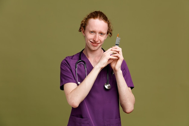 Smiling young male doctor wearing uniform with stethoscope holding syringe isolated on green background
