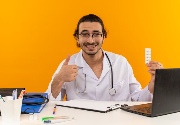 Улыбающийся молодой мужчина-врач в медицинских очках, одетый в медицинский халат со стетоскопом, сидит за столом