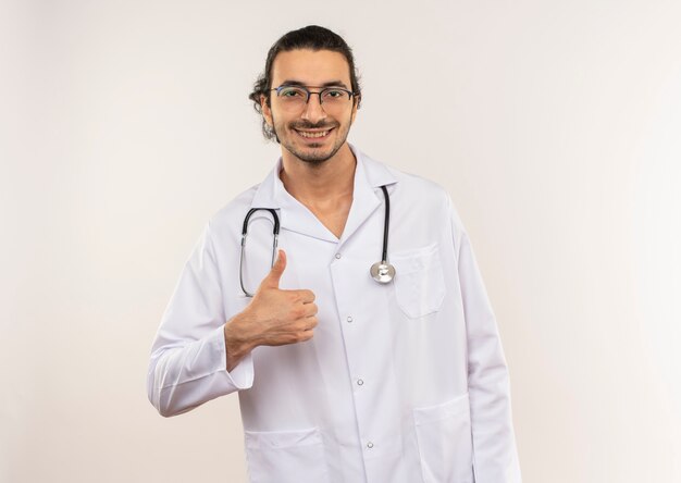 Smiling young male doctor with optical glasses wearing white robe with stethoscope his thumb up on isolated white wall with copy space