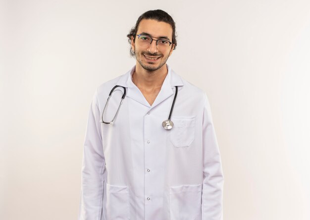 Smiling young male doctor with optical glasses wearing white robe with stethoscope on isolated white wall with copy space