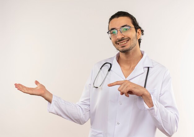 Smiling young male doctor with optical glasses wearing white robe with stethoscope points to side on isolated white wall with copy space