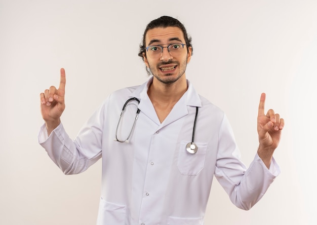 Smiling young male doctor with optical glasses wearing white robe with stethoscope points to up on isolated white wall with copy space