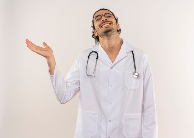 복사 공간이 격리 된 흰 벽에 손으로 청진기 포인트와 흰 가운을 입고 광학 안경 웃는 젊은 남성 의사