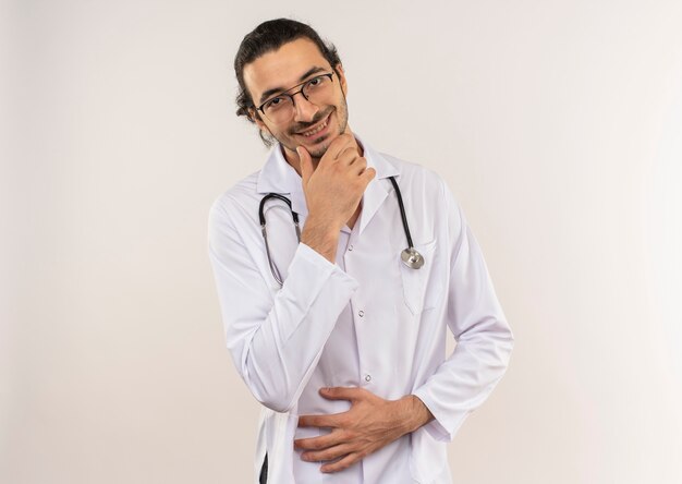 Smiling young male doctor with optical glasses wearing white robe with stethoscope putting hand on chin on isolated white wall with copy space