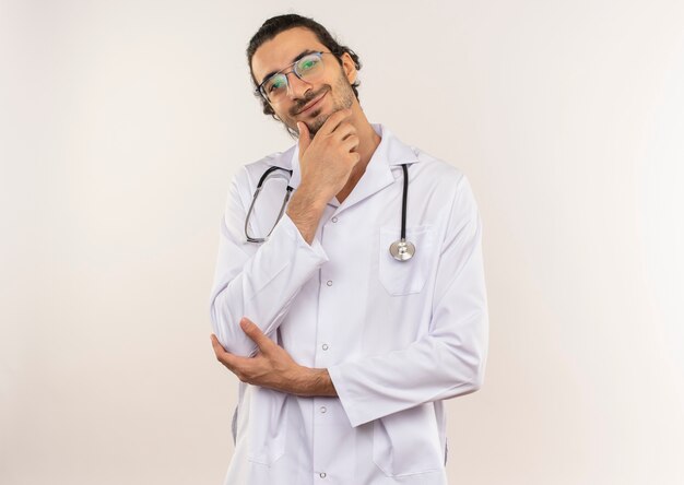 Smiling young male doctor with optical glasses wearing white robe with stethoscope putting hand on chin on isolated white wall with copy space