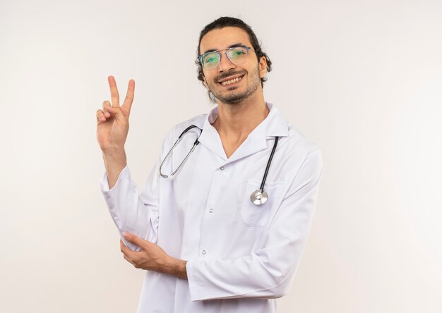 Smiling young male doctor with optical glasses wearing white robe with stethoscope showing peace gesture on isolated white wall with copy space