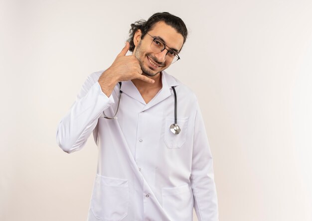 Smiling young male doctor with optical glasses wearing white robe with stethoscope showing phone call gesture on isolated white wall with copy space