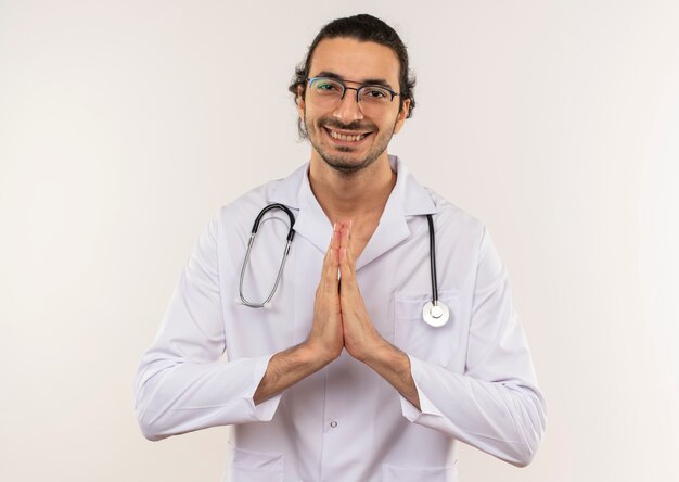 Smiling young male doctor with optical glasses wearing white robe with stethoscope showing pray gesture on isolated white wall with copy space