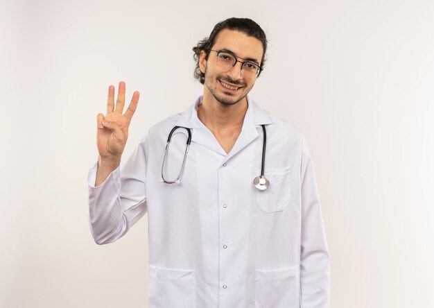 Smiling young male doctor with optical glasses wearing white robe with stethoscope showing three on isolated white wall with copy space