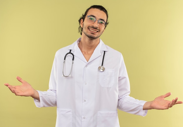 Smiling young male doctor with optical glasses wearing white robe with stethoscope spreads hands on isolated green wall with copy space