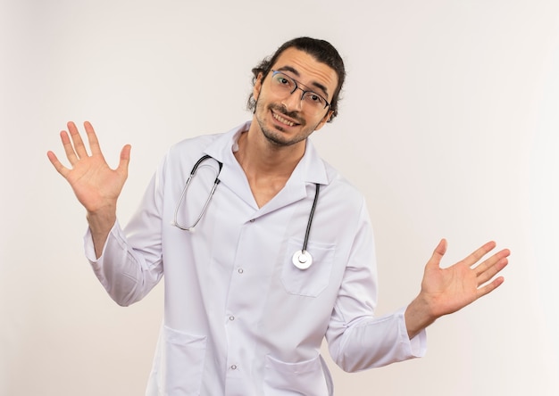 Smiling young male doctor with optical glasses wearing white robe with stethoscope spreads hands on isolated white wall with copy space