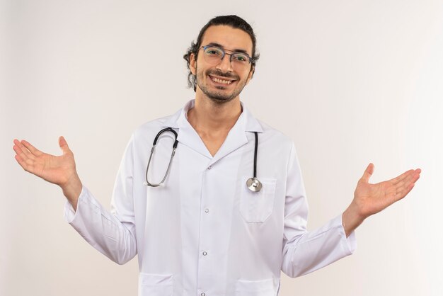 Smiling young male doctor with optical glasses wearing white robe with stethoscope spreads hands on isolated white wall with copy space