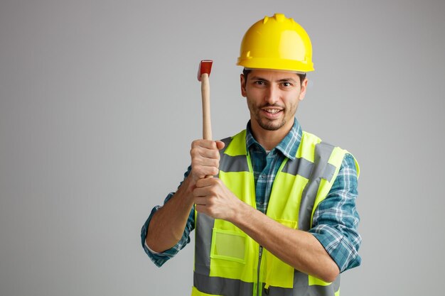 Smiling young male engineer wearing safety helmet and uniform holding hammer looking at camera isolated on white background with copy space