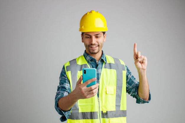 Улыбающийся молодой инженер-мужчина в защитном шлеме и униформе держит и смотрит на мобильный телефон, указывающий вверх на белом фоне