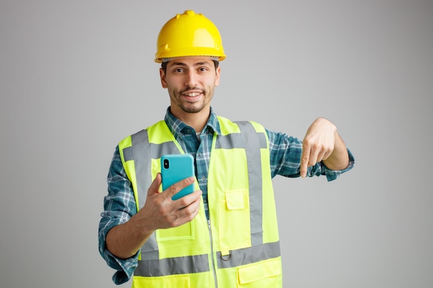 Smiling young male engineer wearing safety helmet and uniform holding mobile phone looking at camera pointing down isolated on white background